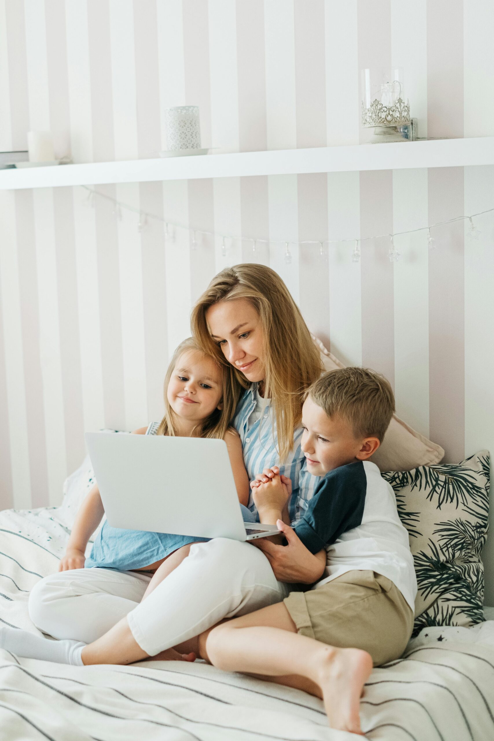 mom sitting on a bed working on a laptop with a little boy and a little girl with her